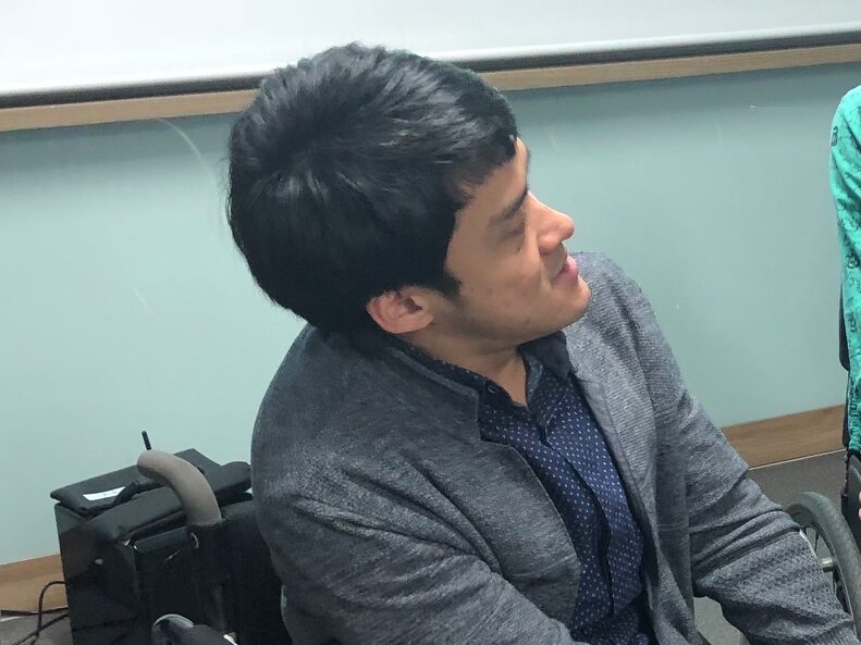 Won Young Kim is looking to their right side as in conversation. They have short black hair and wearing a grey jacket and dark blue shirt.