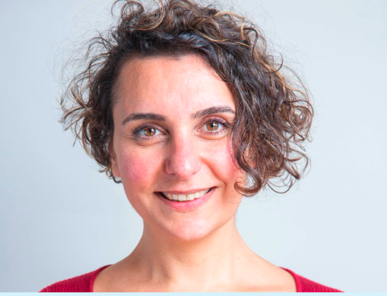 A photograph of Alia Alzougbi smiles directly at the camera with a side curly bob and wearing a red tshirt.