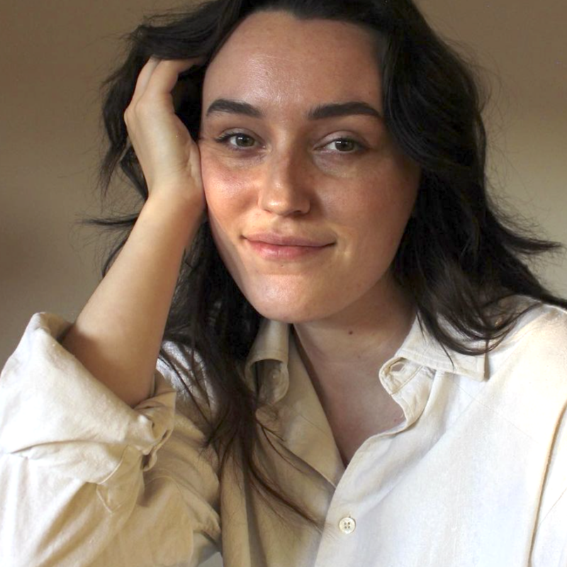 A portrait of Louie staring into the camera. They have long dark hair and a wry smile. Their hand is resting on the side of their face.