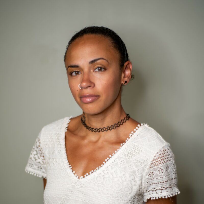 A portrait of Melvee, a mixed race woman wearing a white v neck tshirt. Her hair is tied back and she stands against a grey background.